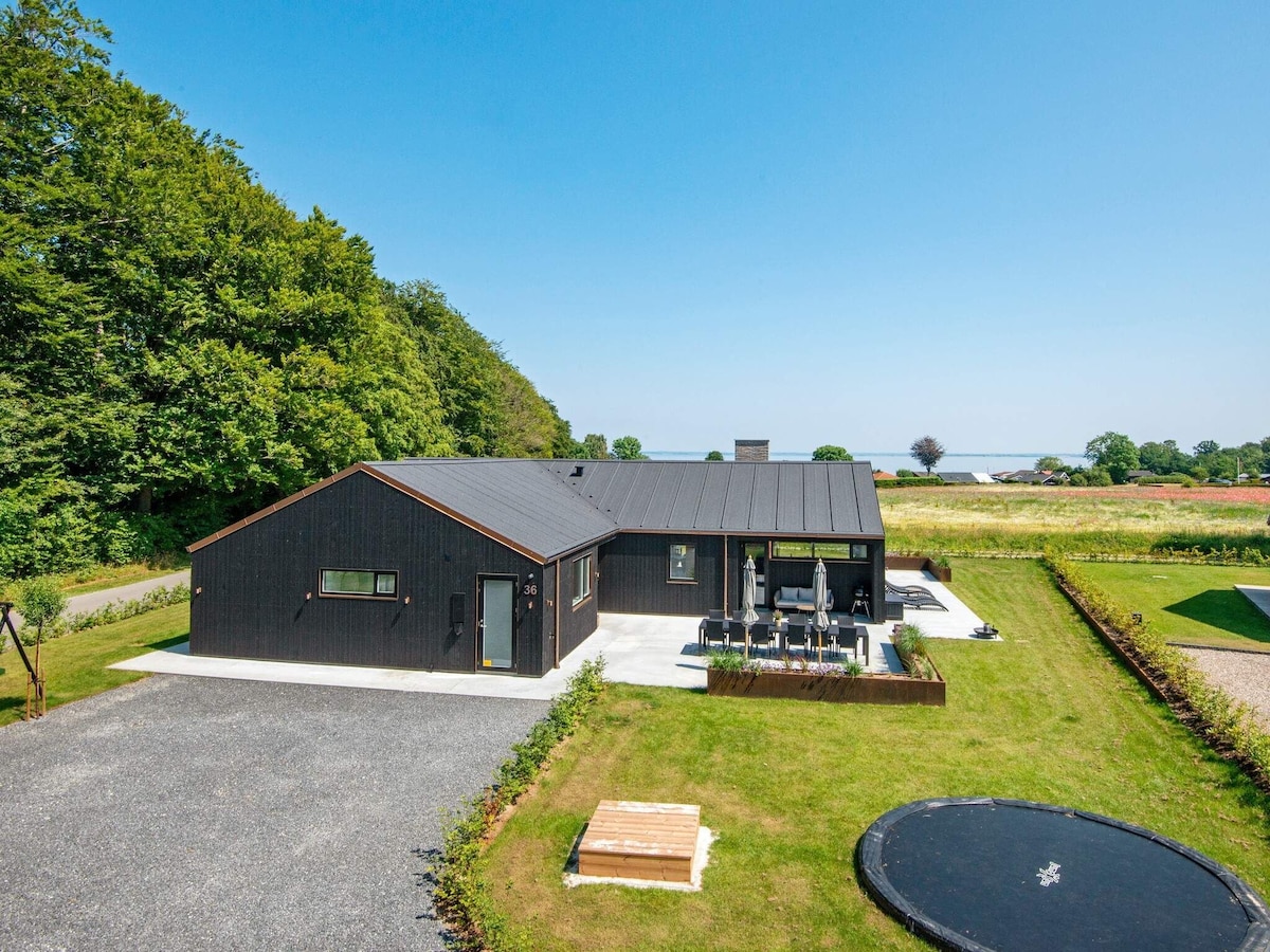 A modern holiday home is nestled in a natural setting, featuring a dark exterior and expansive outdoor areas. A manicured lawn surrounds the property, which includes a trampoline and outdoor seating. The distant horizon offers a glimpse of water under clear blue skies.