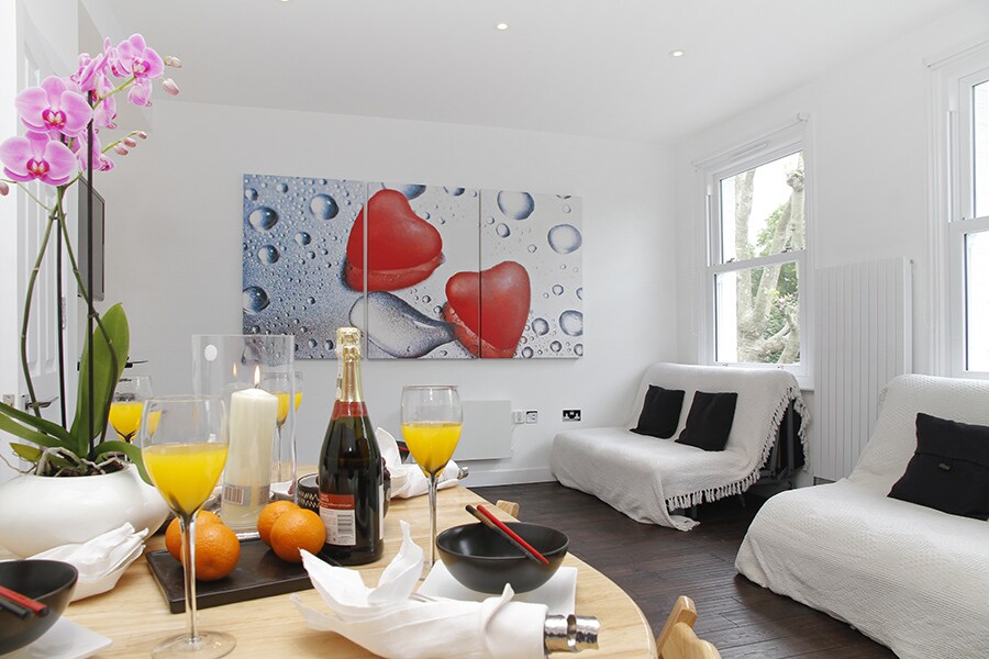 The living area features a round dining table set with glasses of orange juice, accompanied by fruit and elegant tableware. A colorful artwork with red hearts is displayed on the wall. Two sofas draped with white throws are positioned by the window, allowing natural light to enhance the space.