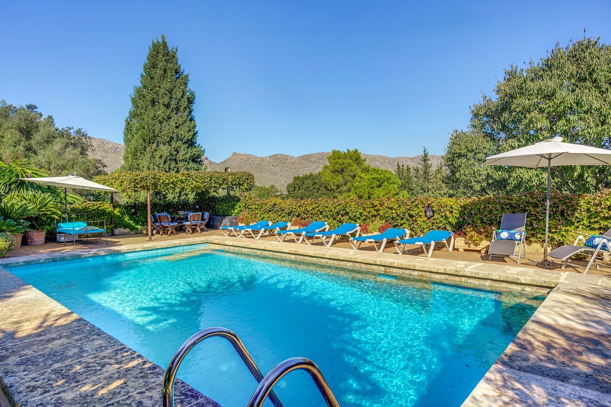 A private swimming pool is surrounded by lush greenery and features sun loungers arranged around its perimeter. Shade is provided by an umbrella near the edge, while a seating area is visible in the background, overlooking the tranquil landscape.