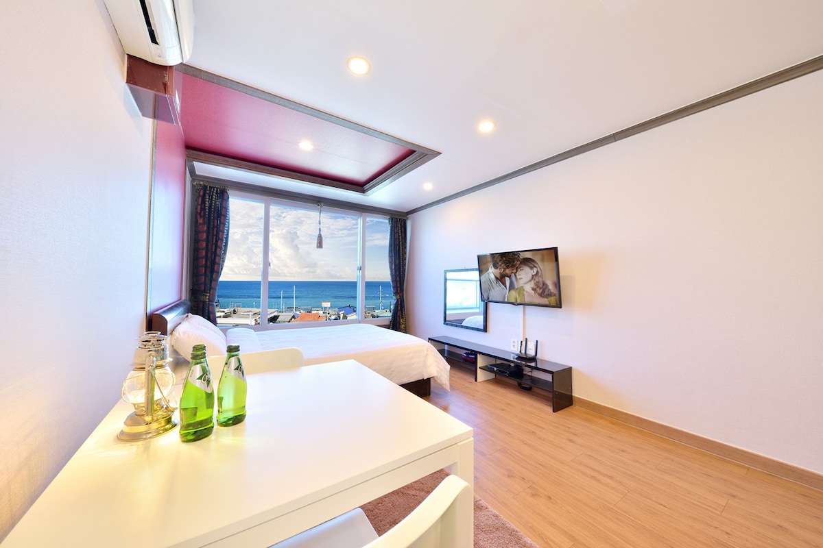 A well-lit room features a spacious bed next to large windows showcasing a view of the ocean. A small dining table is set with bottles of water, and a flat-screen TV is mounted on the wall, providing modern amenities within a calming space.