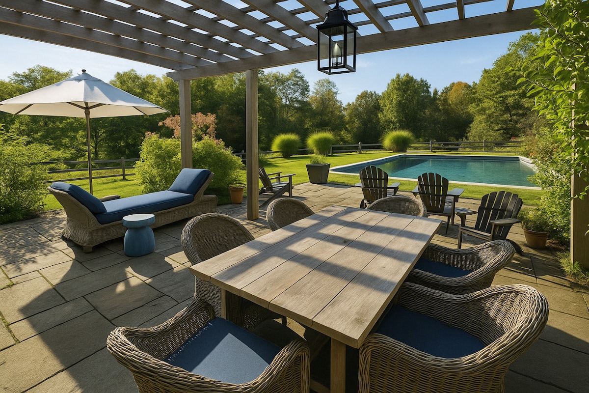 A shaded outdoor patio is highlighted by a wooden dining table surrounded by comfortable chairs. A lounge chair and umbrella offer relaxation options, while an in-ground pool glimmers in the background, framed by lush greenery and scenic views.