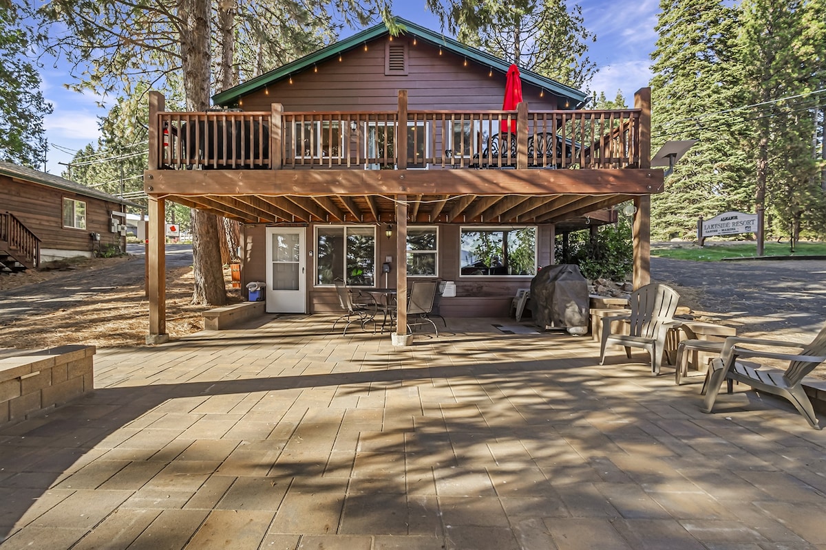 A wooden deck extends from the cabin, offering ample space for seating. The patio area is paved, featuring several chairs and a table, surrounded by trees and natural landscaping. Sunlight casts playful shadows across the ground, creating a warm atmosphere.
