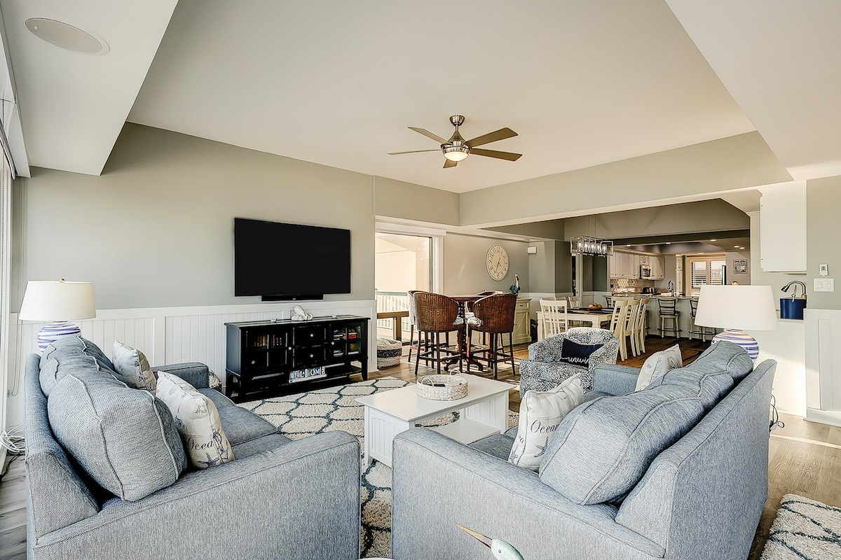 A spacious living area showcases two comfortable gray sofas positioned around a coffee table, with a ceiling fan overhead. A wall-mounted TV is visible, and the open layout leads to the dining area and modern kitchen in the background, enhancing the sense of space and connection.