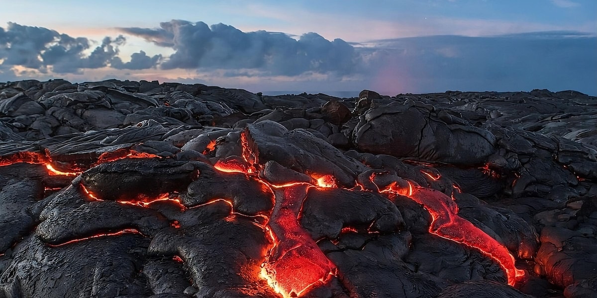 A vast surface of hardened lava is visible, with bright orange lava peeking through cracks and crevices. The landscape appears rugged and textured, under a sky filled with clouds illuminated by the sunset, showcasing a range of colors.