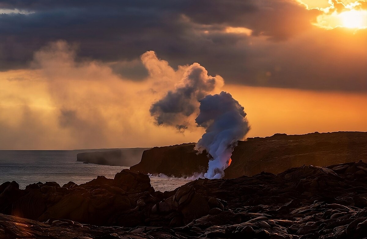 A dramatic view of steam rising from the ocean meets lava at the shoreline during sunset. The smoke contrasts against the warm hues of the sky, while dark volcanic rock frames the lower portion of the image, emphasizing the dynamic landscape.