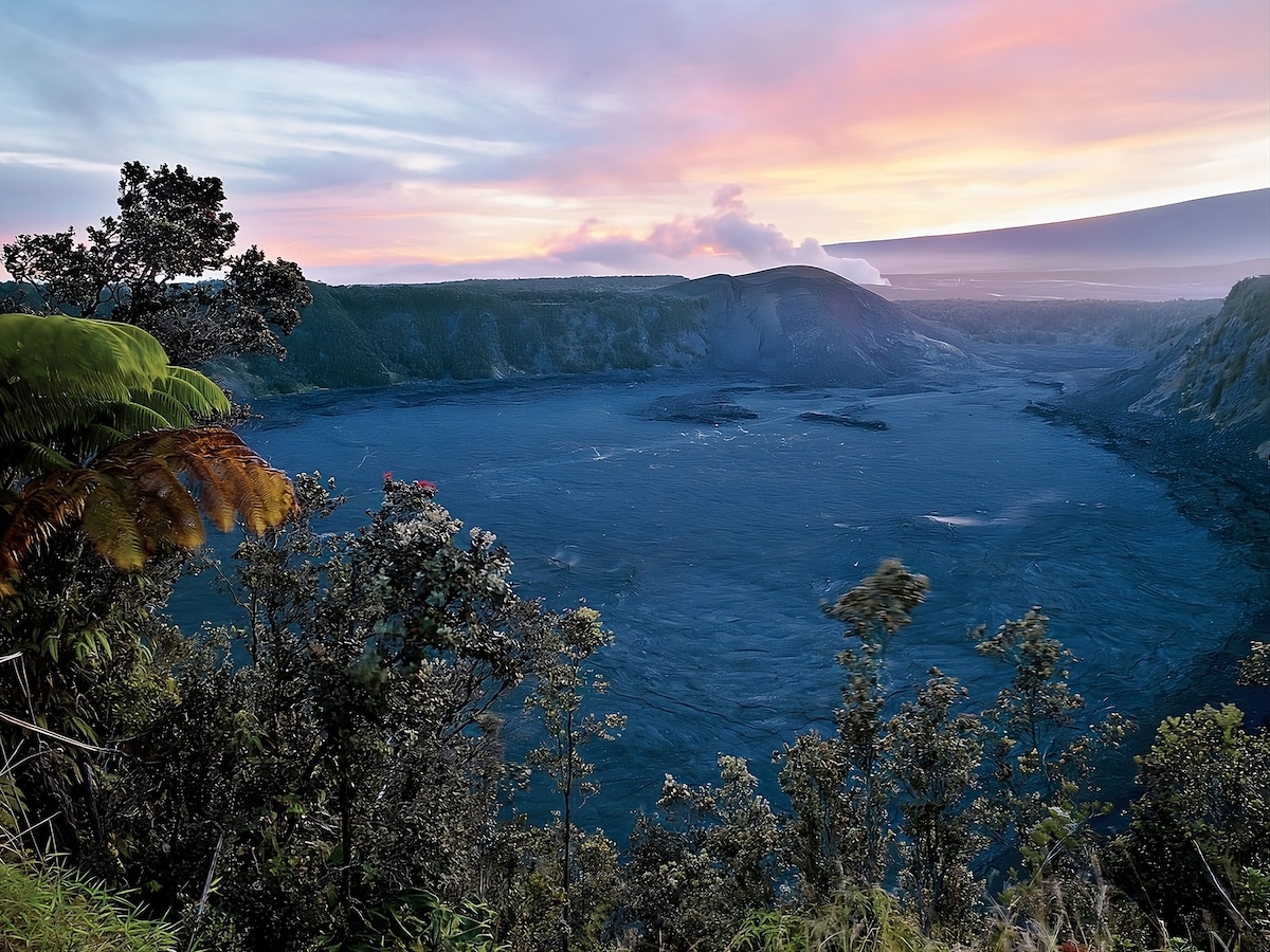 A panoramic view of a volcanic crater is showcased, with a blue-grey landscape emerging from surrounding greenery. The sky reflects hues of pastel pink and purple during sunset, while clouds rest over the crater's edge, creating a serene connection to the natural environment.