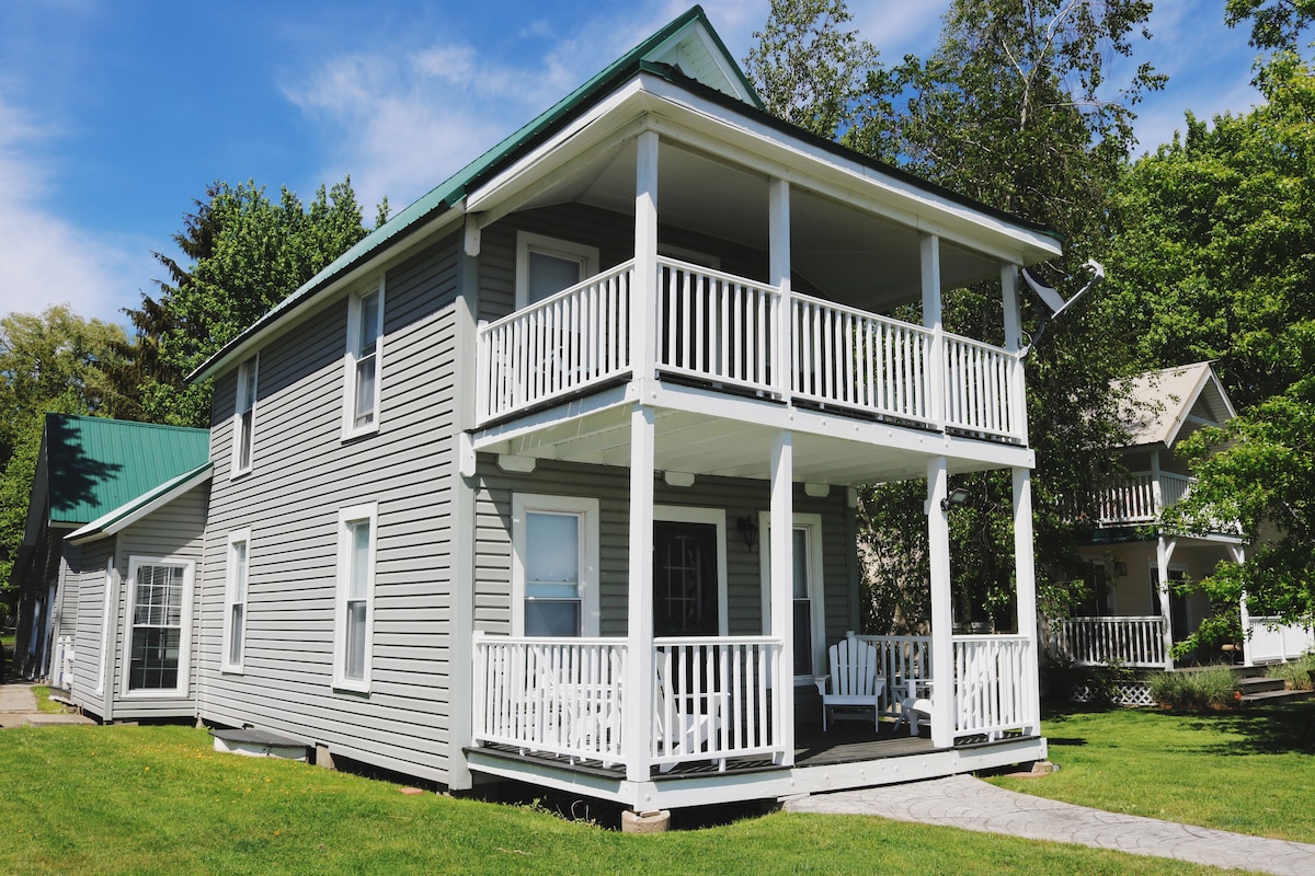 The exterior of a two-story cottage is presented, featuring a light grey facade and white railings. Two covered decks are visible, with chairs on the lower level. Lush green grass surrounds the property, while trees can be seen in the background, indicating a peaceful environment.