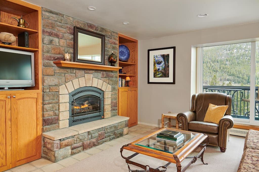 A living area features a stone fireplace flanked by wooden bookshelves. A comfortable armchair is positioned nearby, with a large window providing views of the surrounding mountains. Decorative accents, including a framed artwork and a coffee table, complete the inviting space.
