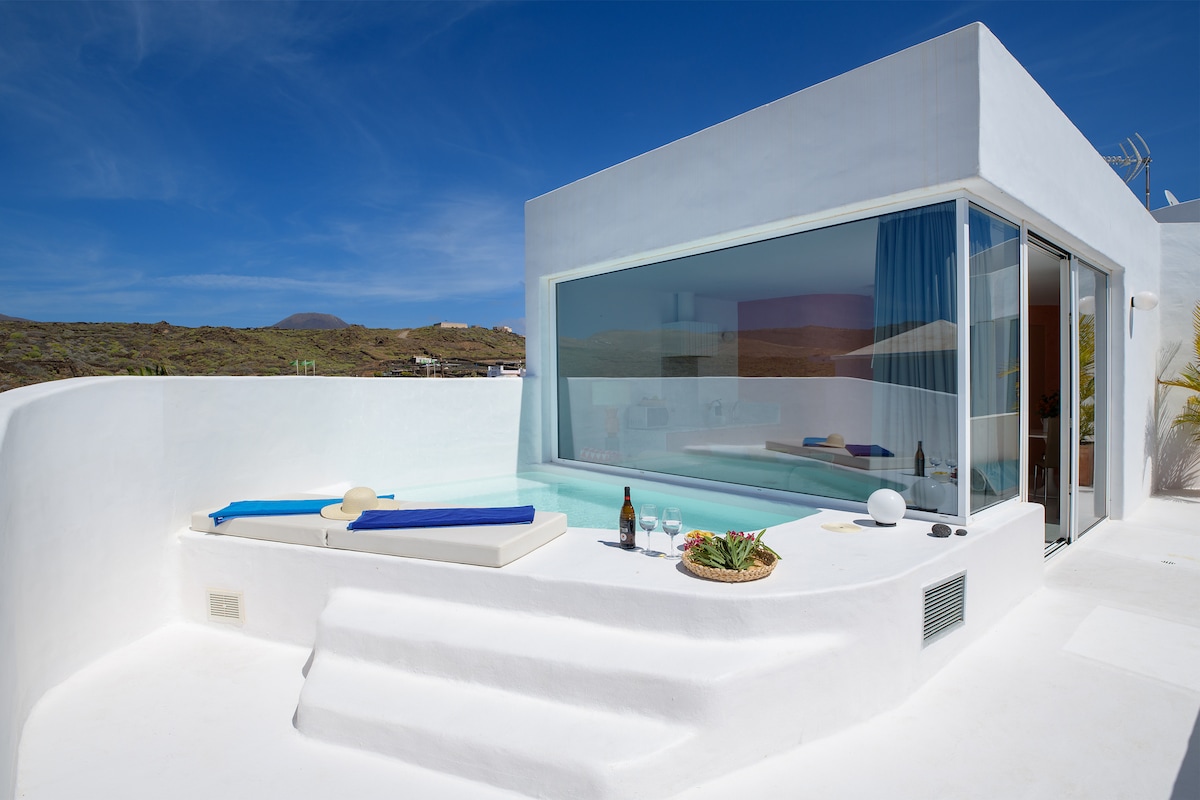 A bright private terrace features a small pool surrounded by lounge chairs. Large glass windows provide unobstructed views of the landscape. The outdoor area is designed with clean lines and minimal decor, creating a serene space for relaxation.