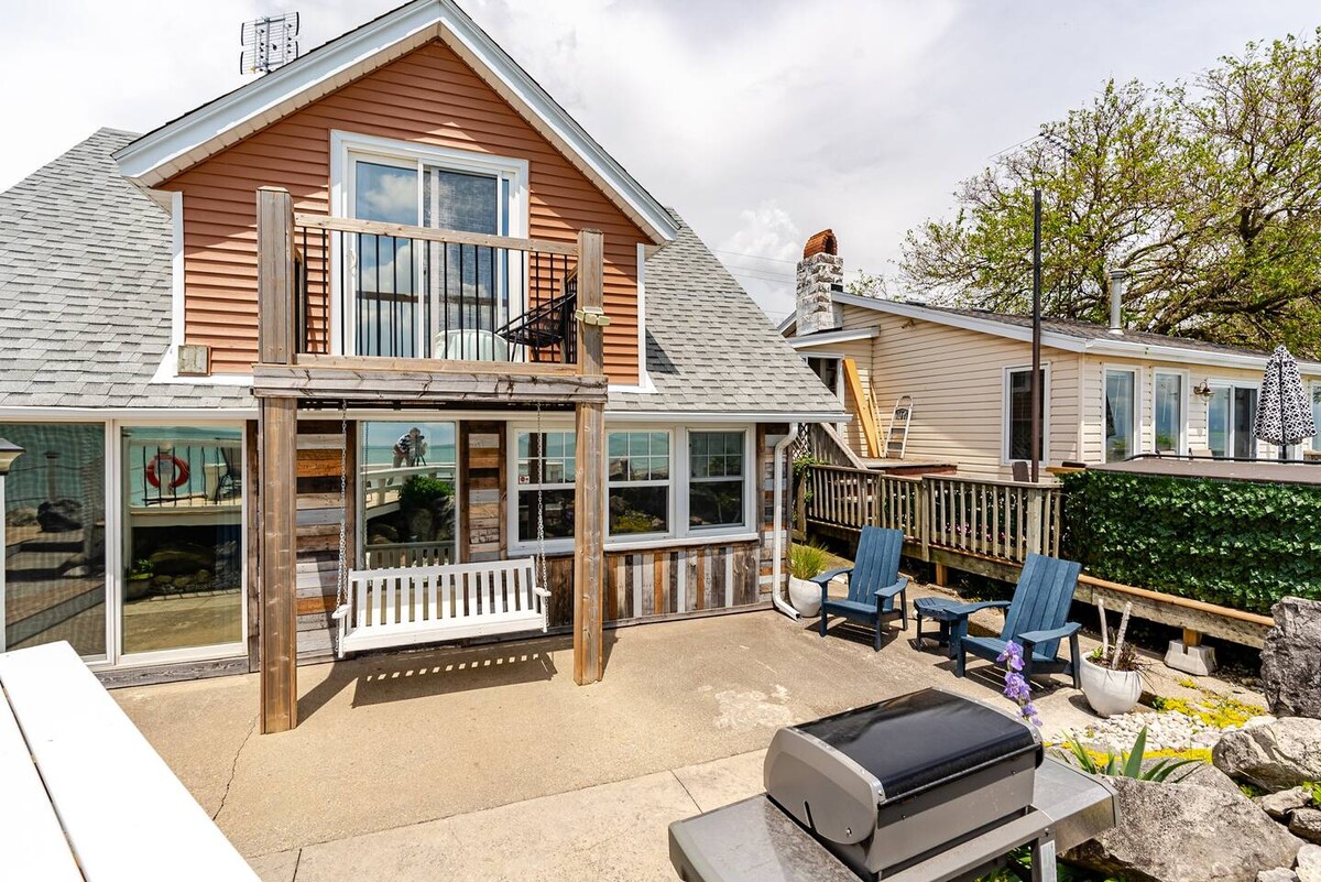 The exterior of the cottage showcases a welcoming waterfront patio, featuring comfortable chairs and a swing. The second-floor balcony offers a view of the surroundings. A propane BBQ grill is visible, with a backdrop of trees and nearby structures, creating an inviting outdoor space.
