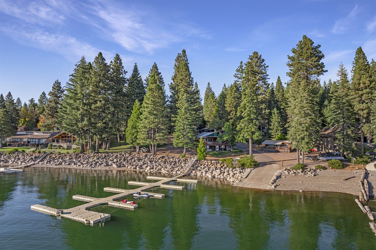 A serene lakeside view showcases a collection of docks extending into Lake Almanor, surrounded by lush pine trees. The property features a spacious lawn leading to the water, where rocky shoreline complements the natural landscape. The inviting blue water reflects the clear sky.