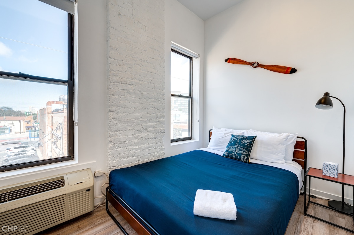 A bright sleeping area features a spacious bed with a navy blue coverlet and a decorative pillow. A white towel is neatly placed at the foot of the bed. Natural light filters in through two windows, complemented by a modern floor lamp and a vintage airplane propeller accent above the bed.