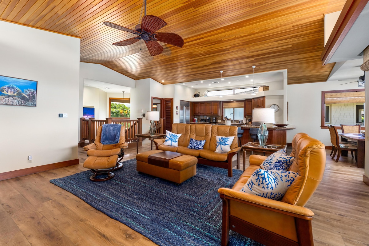 A spacious living area features comfortable leather seating arranged around a central coffee table, with decorative pillows adding a touch of colour. A ceiling fan hangs overhead, and wooden beams adorn the ceiling, enhancing the open atmosphere. The layout flows seamlessly into the dining and kitchen spaces.