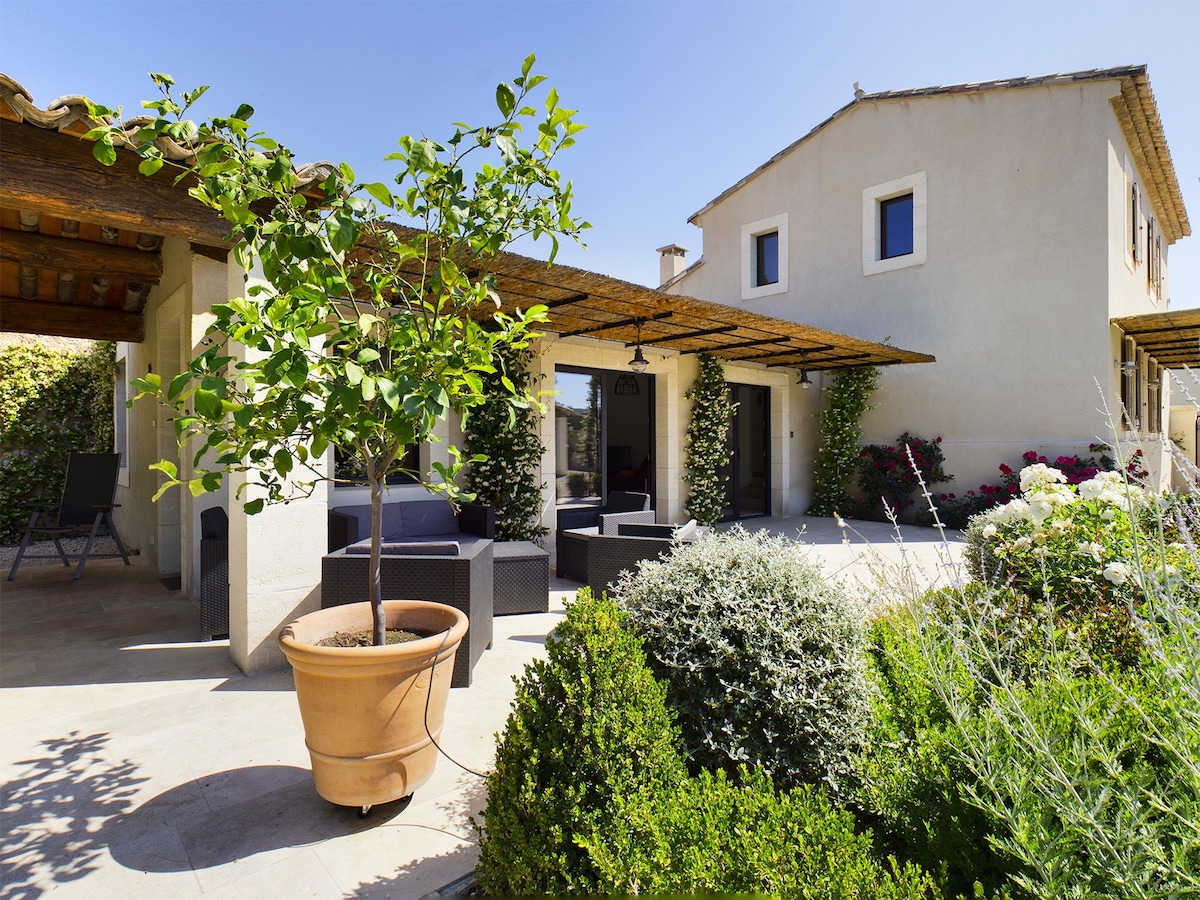 The outdoor area features a terracotta planter with lush greenery, surrounded by well-maintained shrubs and flowers. Comfortable seating areas are positioned beneath wooden beams, with the villa's light-colored exterior and adjoining garden visible in the background.