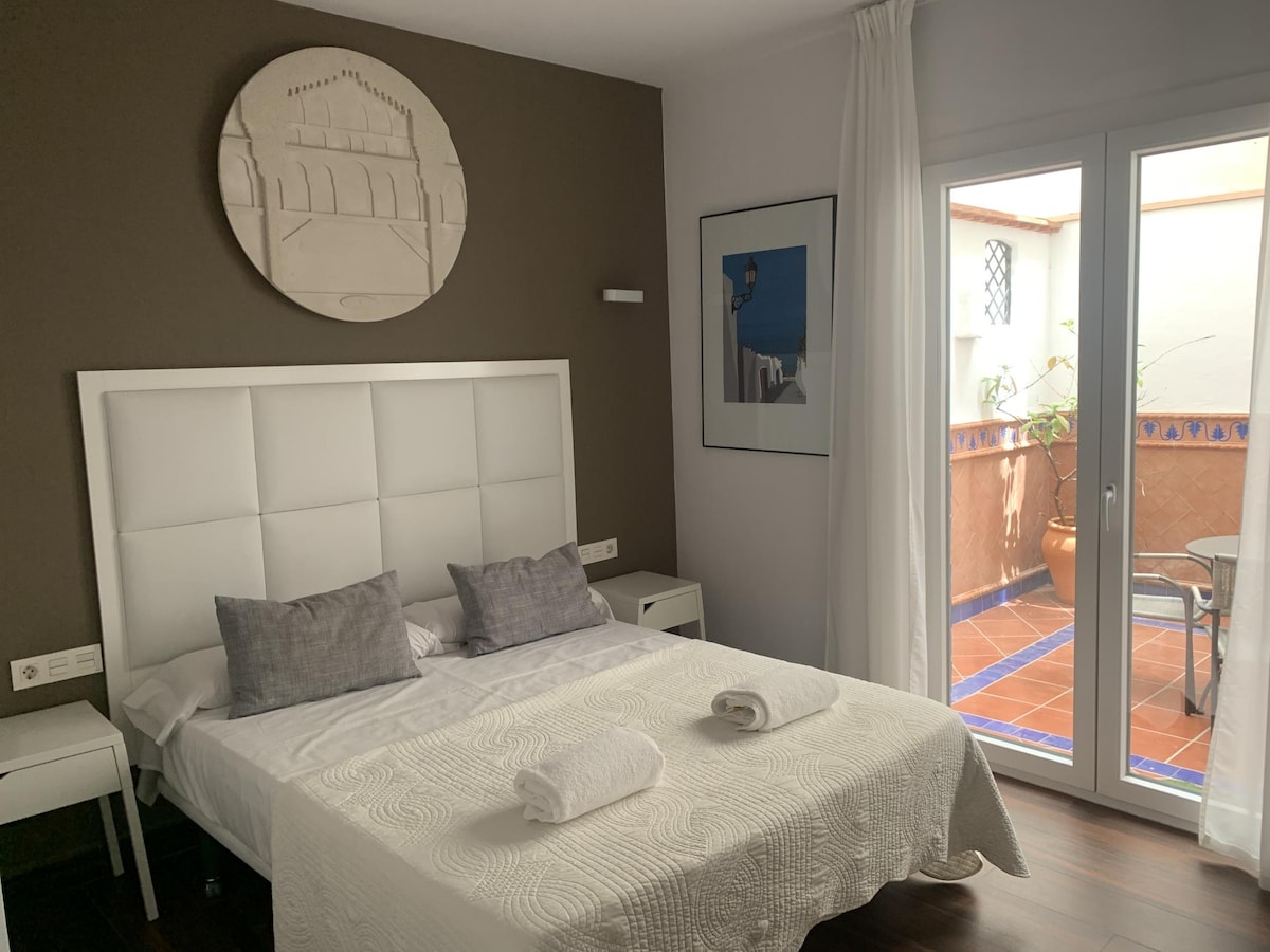 A minimalist bedroom features a spacious bed with light bedding and two gray pillows. A large piece of art adorns the wall above the bed. Natural light enters through glass doors leading to a private terrace, enhancing the calm environment.