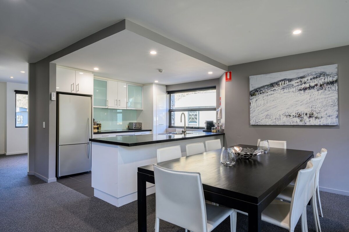 A modern kitchen and dining area features sleek cabinetry and a spacious island counter. A large dining table with white chairs accommodates multiple guests, while natural light streams through a large window. A contemporary piece of artwork adorns the wall, complementing the neutral colour scheme.