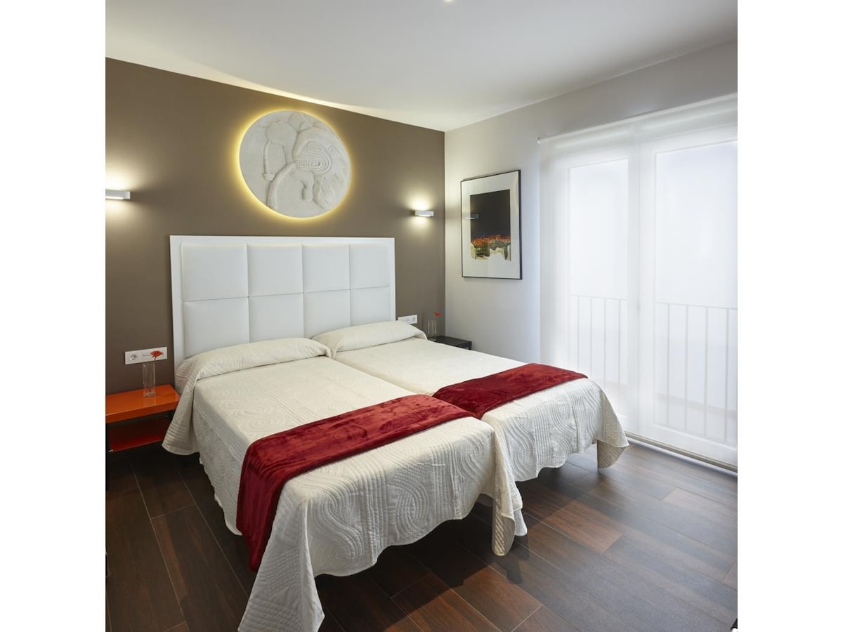 A bright sleeping area features two twin beds covered with light-colored bedding and topped with deep red throws. A decorative wall relief provides visual interest, while natural light enters through a set of glass doors leading to a small balcony.