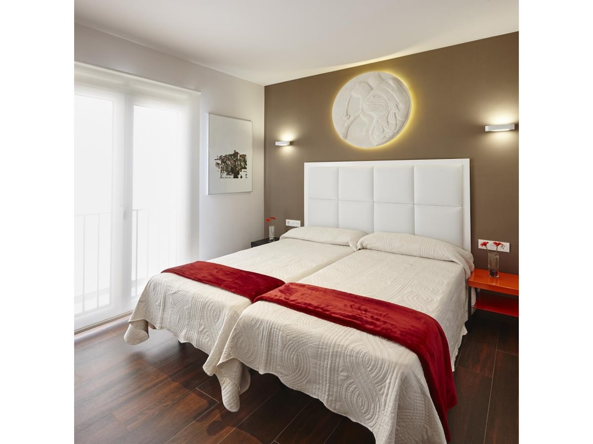 A sleeping area features two spacious beds dressed in neutral-toned linens, complemented by red throw blankets. Wall sconces provide soft lighting, while a decorative wall panel adds a unique touch. Large glass doors lead to a small balcony, allowing natural light to fill the room.