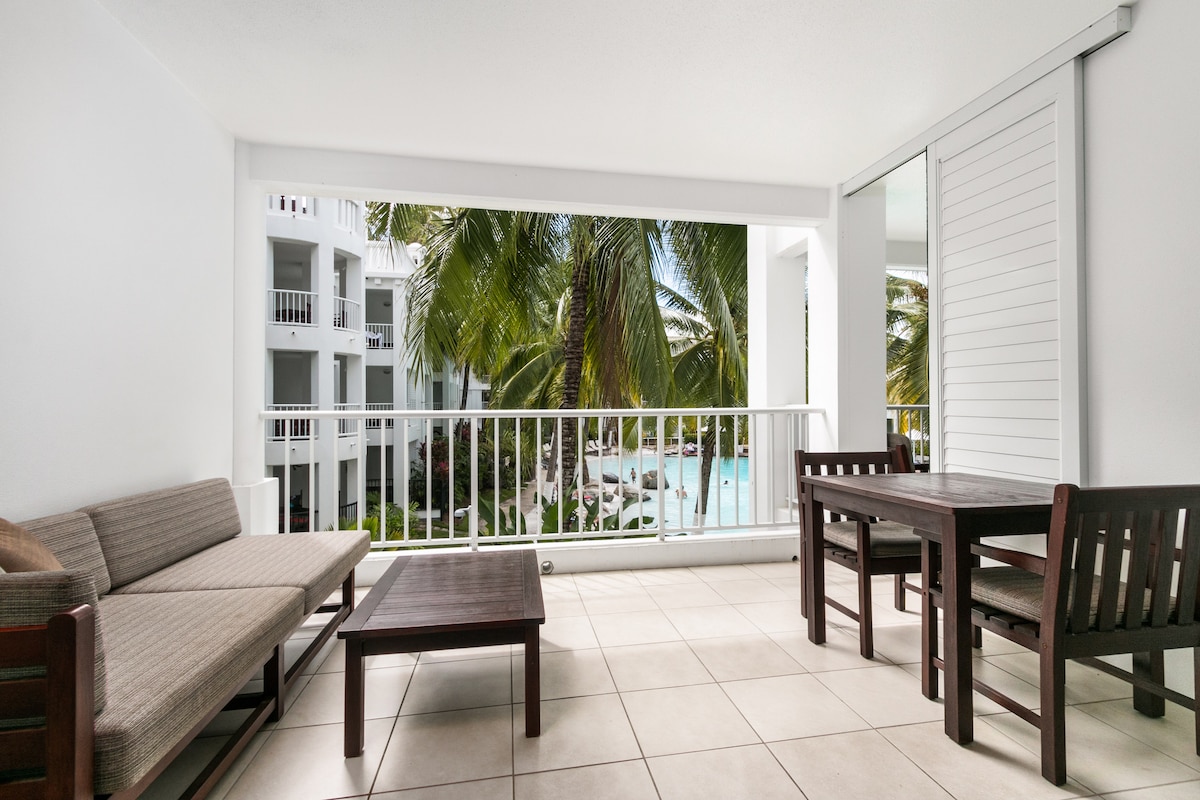 A spacious balcony is highlighted by modern outdoor furniture, including a sofa, coffee table, and dining set. Lush palm trees are visible in the background, framing the views of the Lagoon Pool. Large sliding doors allow natural light to fill the space.