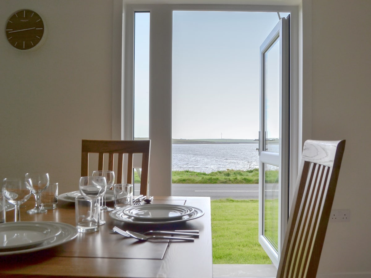 An open door reveals scenic views of the coastline, allowing natural light to brighten the dining area. A wooden table is set with plates and glassware, ready for a meal, while the distant water can be seen beyond the grassy foreground.