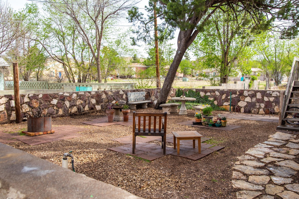 A shaded outdoor area features a rustic stone wall and several seating arrangements, including wooden benches and chairs. Potted plants are placed on stone surfaces, with a tree providing natural shade. The ground consists of gravel and pavers, creating an inviting space for relaxation.