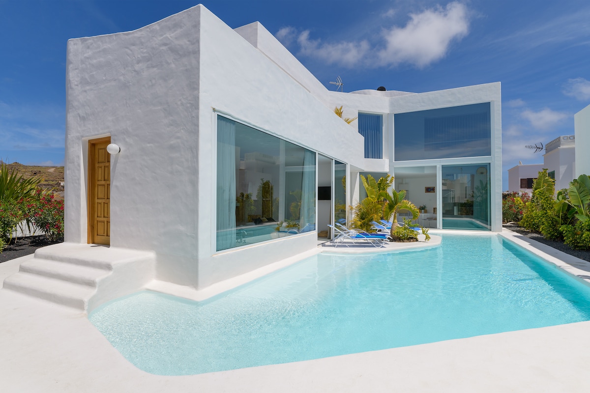 A modern building features large glass windows and a spacious swimming pool. The pool reflects the clear blue sky and is surrounded by lush greenery. The entrance is marked by a wooden door, adding a touch of warmth to the contemporary design.