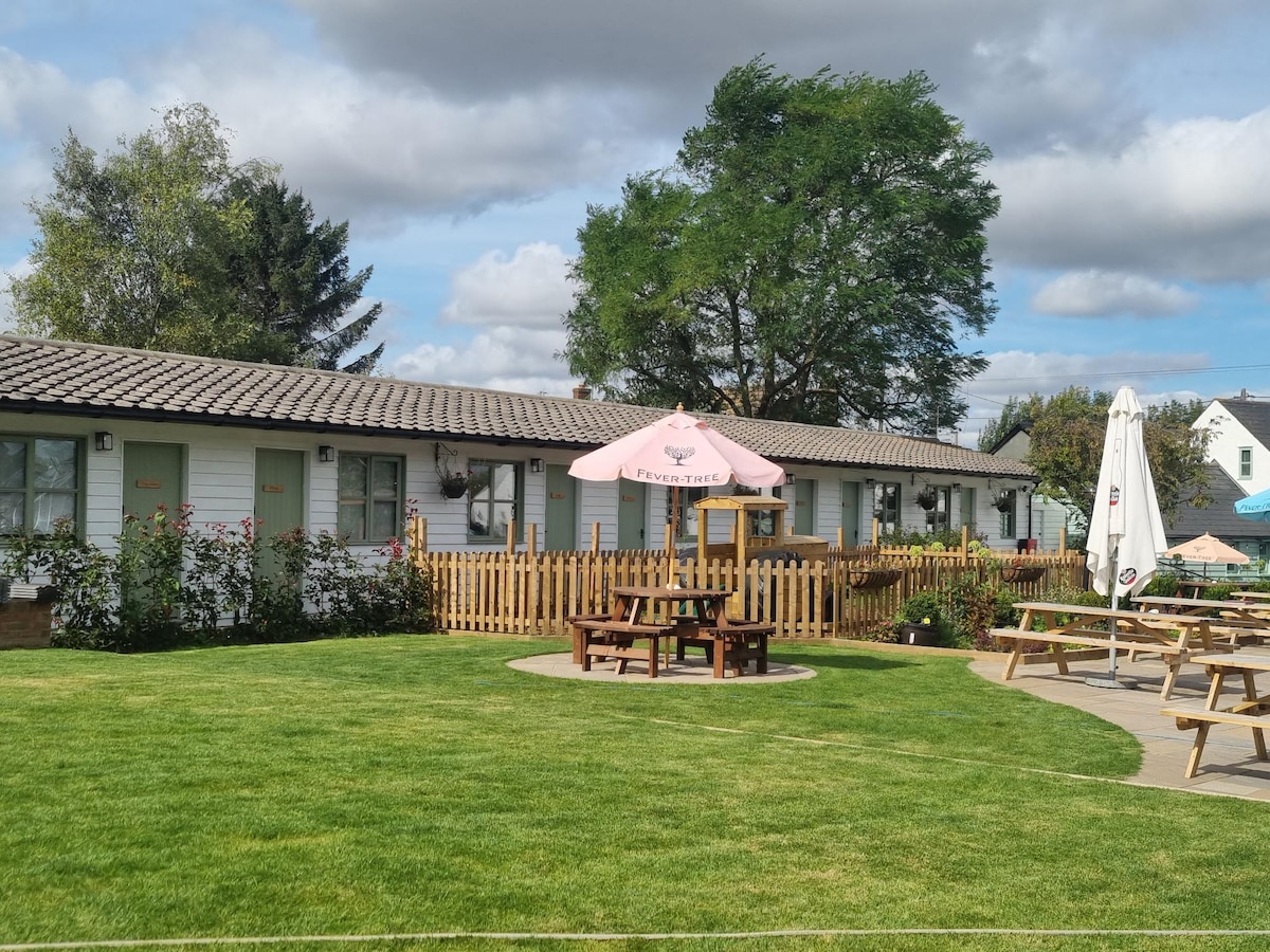 A cluster of self-contained chalets is presented, framed by a well-maintained lawn. Wooden picnic tables are placed under umbrellas, inviting relaxation. Green trees provide shade, while a wooden fence surrounds the area, contributing to the tranquil ambiance of the countryside setting.