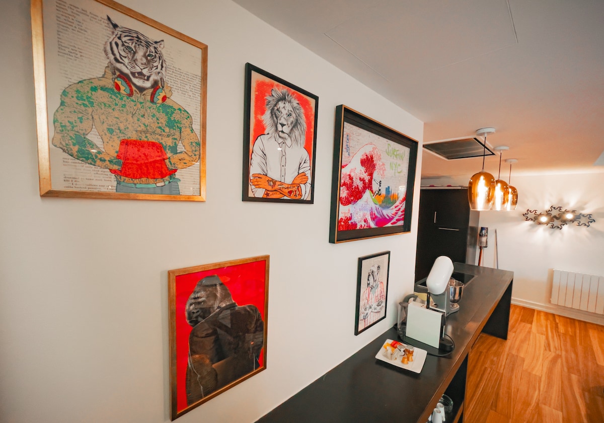 A kitchen area features a dark countertop complemented by a collection of framed artwork on the wall. The pieces include vibrant illustrations of animals and abstract designs. Warm pendant lights hang above, enhancing the modern aesthetic of the space.