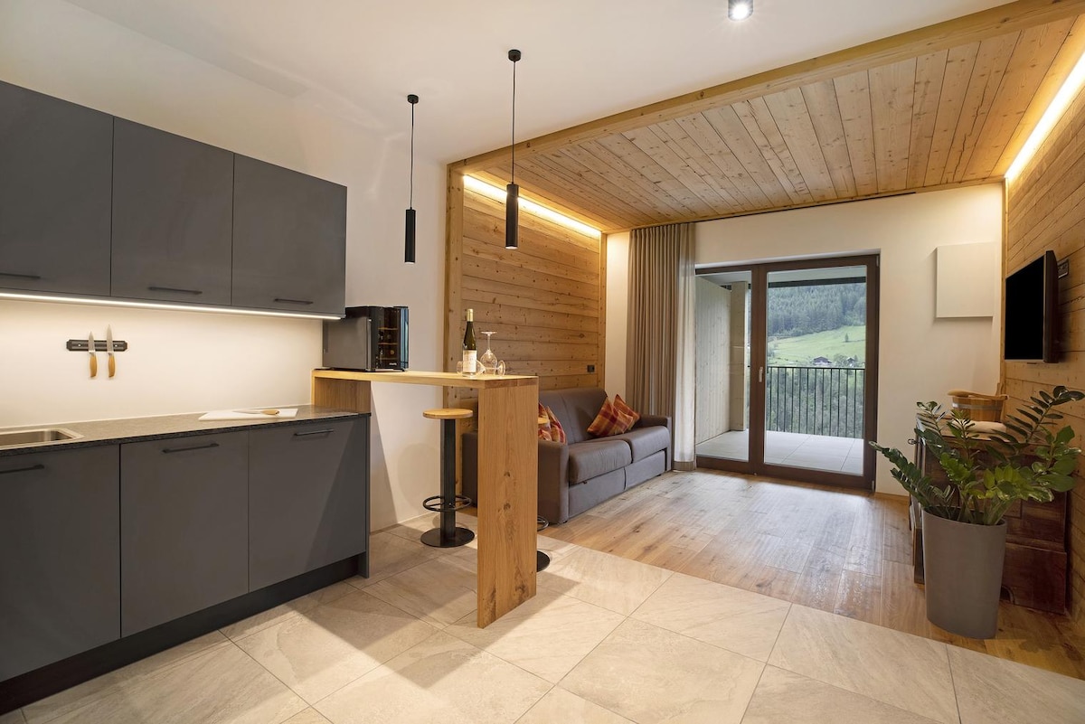 A modern kitchen area is equipped with dark cabinetry and integrated appliances, featuring a countertop that transitions into a small dining space. A cozy sofa sits adjacent to the sliding glass doors, leading to a private balcony with views of the surrounding landscape.