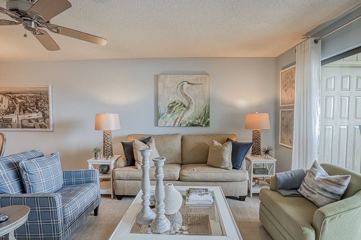 A light and airy living room is presented, featuring a neutral-toned sofa complemented by a patterned accent chair. Table lamps add warmth to the space, while a decorative painting of a heron adorns the wall. Natural light floods in through a nearby doorway.