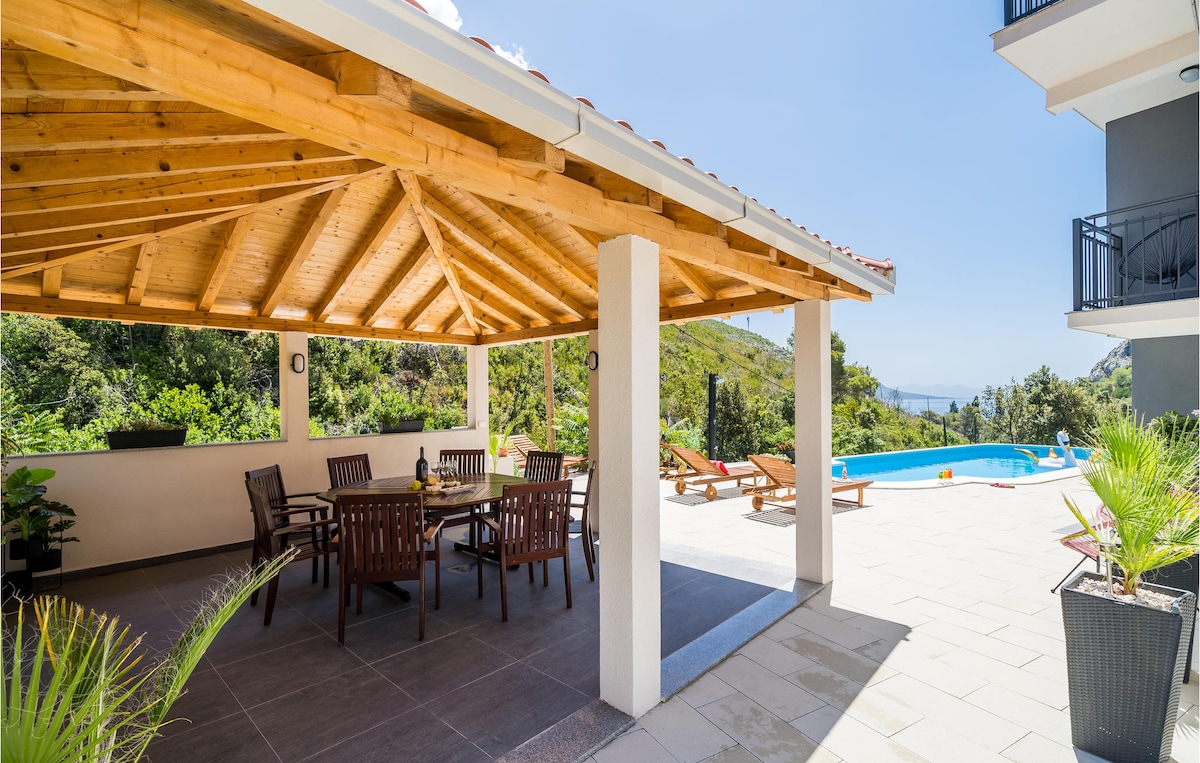 A covered terrace features a large dining table surrounded by wooden chairs, providing a shaded area for meals. The area is complemented by views of the pool and surrounding greenery, creating a serene setting for gatherings.