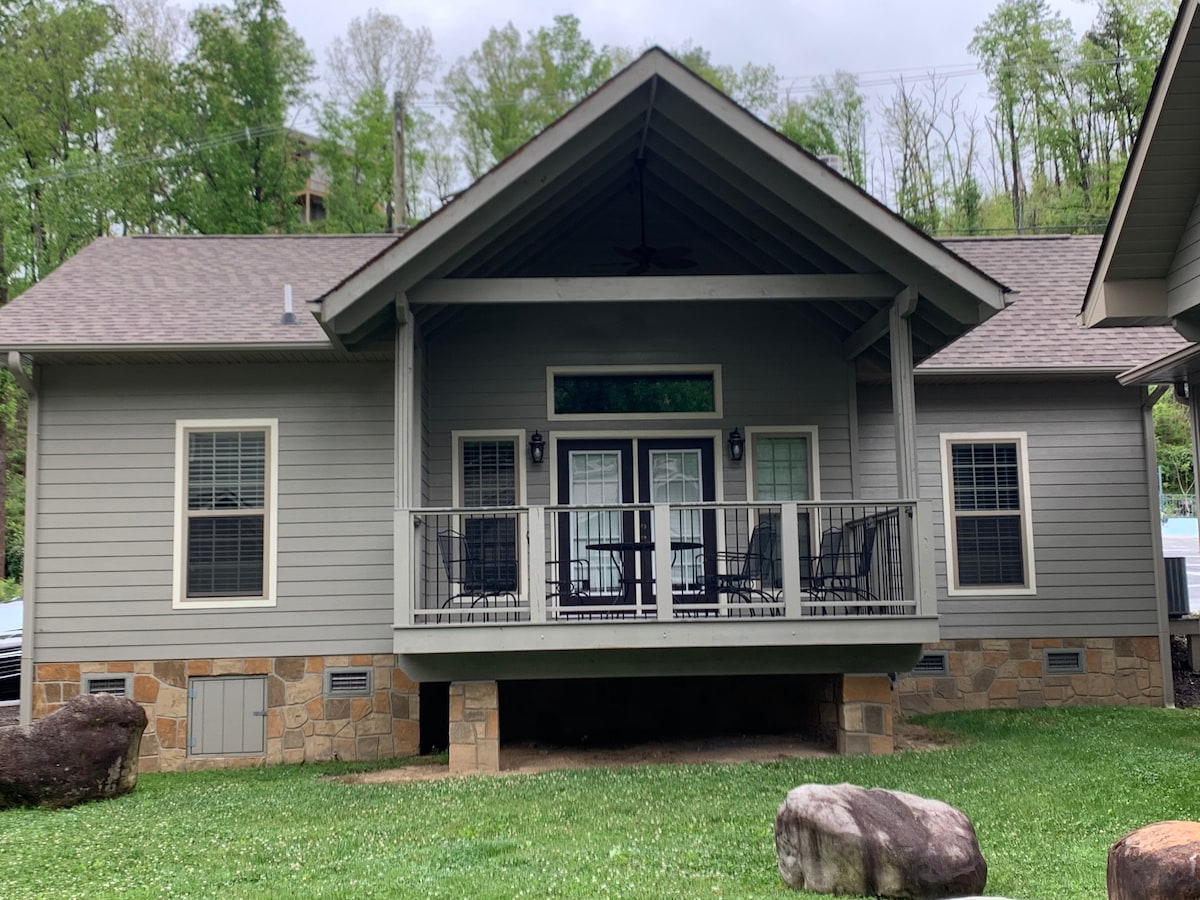 The cottage exterior is highlighted by a broad balcony featuring wooden rocking chairs. The entrance is framed by large glass doors, providing easy access to the interior. Natural stone accents and a green lawn enhance the inviting atmosphere of the surrounding landscape.