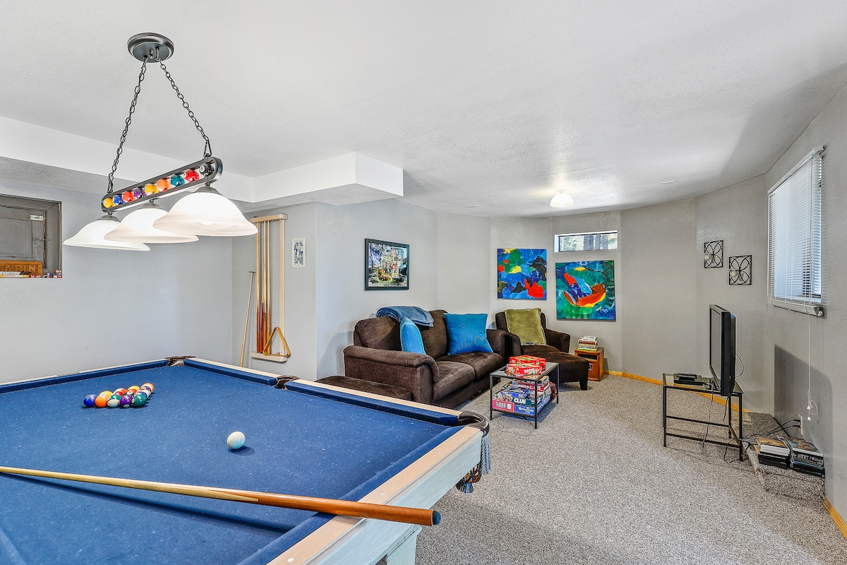 A spacious game room features a pool table in the foreground with colorful balls arranged on the felt. A comfortable seating area with a couch and coffee table is visible, accompanied by wall art on light grey walls and a TV positioned in the corner.