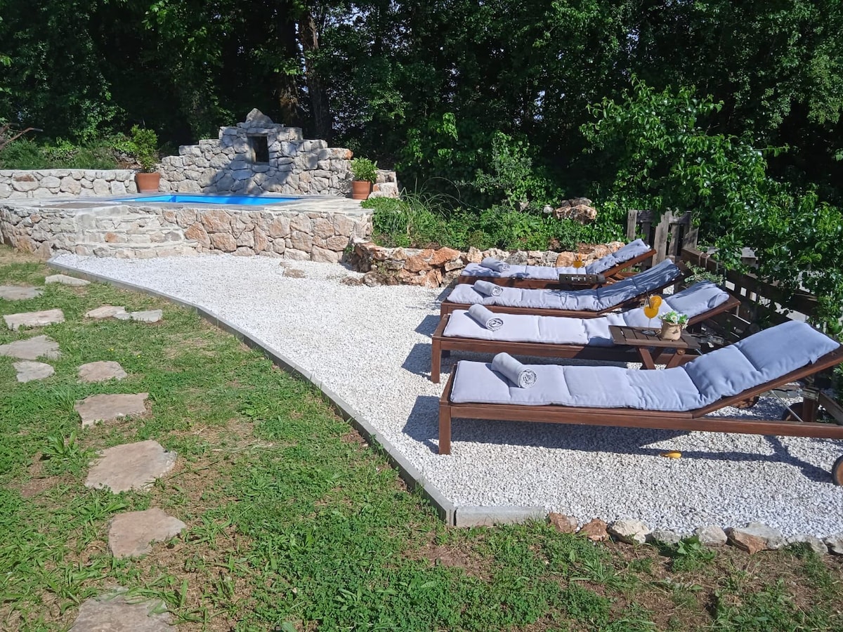 A tranquil outdoor space features several lounge chairs arranged on a smooth gravel area. A stone wall encloses a small pool, surrounded by greenery, providing a relaxing environment for guests to unwind.