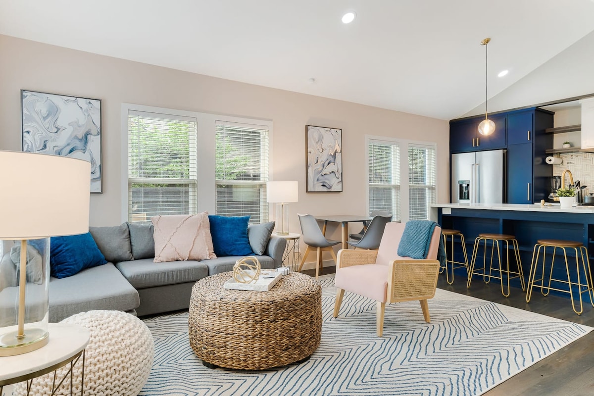 A well-lit living area features a sectional sofa adorned with soft pillows and a round woven coffee table at the center. Six dining chairs surround a kitchen peninsula, showcasing stainless steel appliances and a stylish light fixture overhead. Natural light filters through multiple windows.