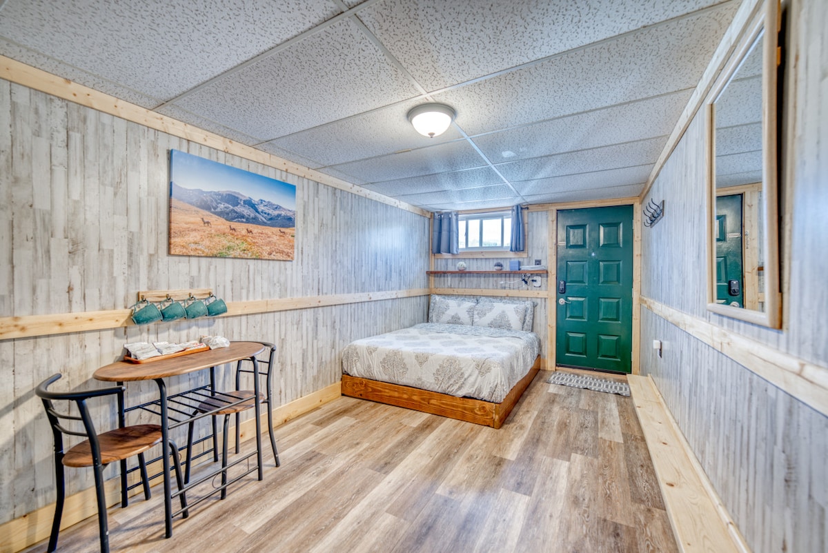 A bright room features warm wood paneling, a spacious bed, and a small dining/work table with two chairs. A mountain landscape artwork decorates the wall, while a door and window provide natural light. The floor is finished in a light wood tone.