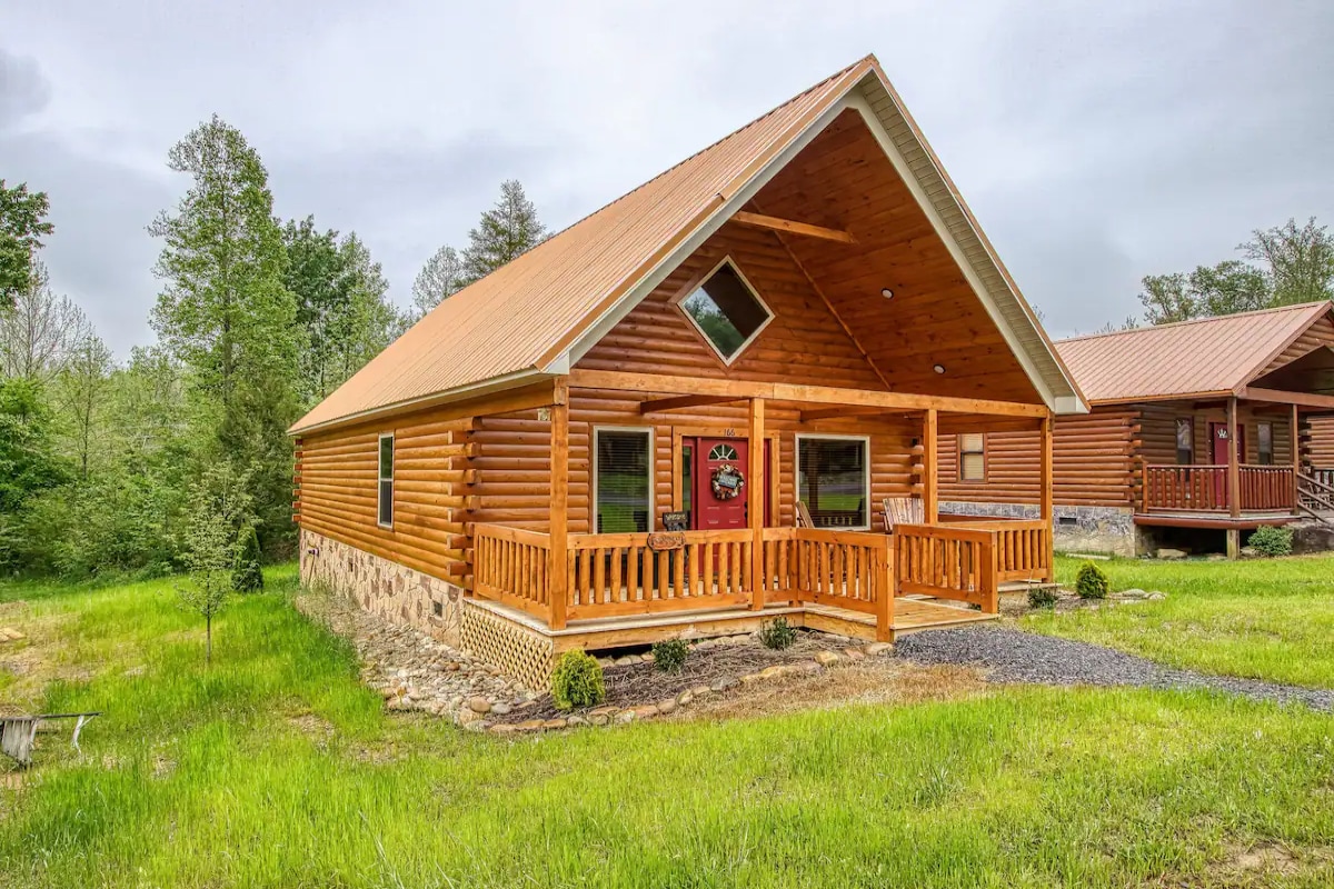 The exterior of a charming log cabin is showcased, featuring a peaked roof and a welcoming front porch. A well-maintained lawn surrounds the property, with green grass and small shrubs enhancing the natural setting. The cabin's rustic charm is complemented by large windows and traditional wood siding.
