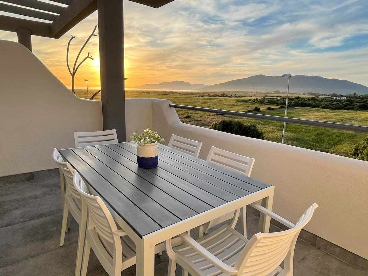 An outdoor terrace is presented, featuring a large dining table with white chairs, set against a backdrop of a sunset illuminating the landscape. Soft hues of orange and yellow fill the sky, while distant mountains create a serene horizon.