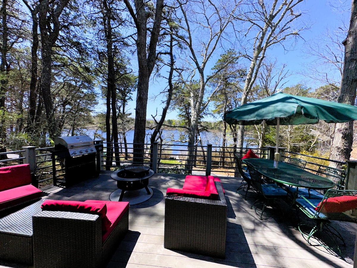 An outdoor deck offers a scenic view of Minister’s Pond, surrounded by tall trees. Comfortable seating with red cushions and a matching firepit is available. A patio table is positioned under a green umbrella, and a gas grill is visible in the background, making it ideal for gatherings.