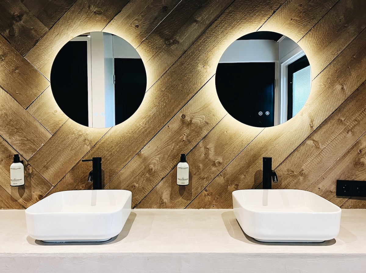 A modern bathroom features two round mirrors framed by ambient lighting above square sinks. The sinks are positioned on a clean countertop, complemented by minimalist soap dispensers mounted on the wall. Natural wood paneling creates a warm backdrop.