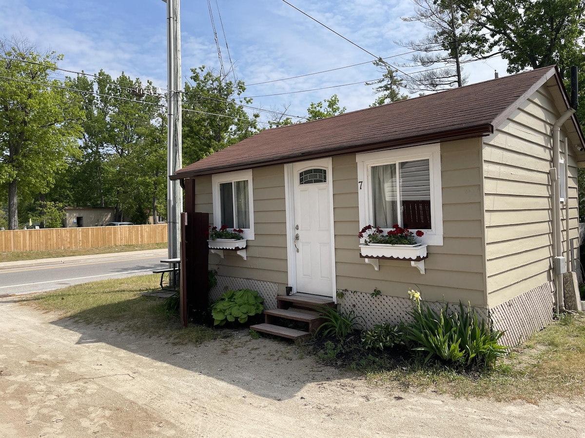 A modest cottage exterior featuring a light brown façade with white trim and flower boxes. The front door leads to a welcoming entrance, and greenery surrounds the base of the building. A gravel path extends towards a nearby street lined with trees.