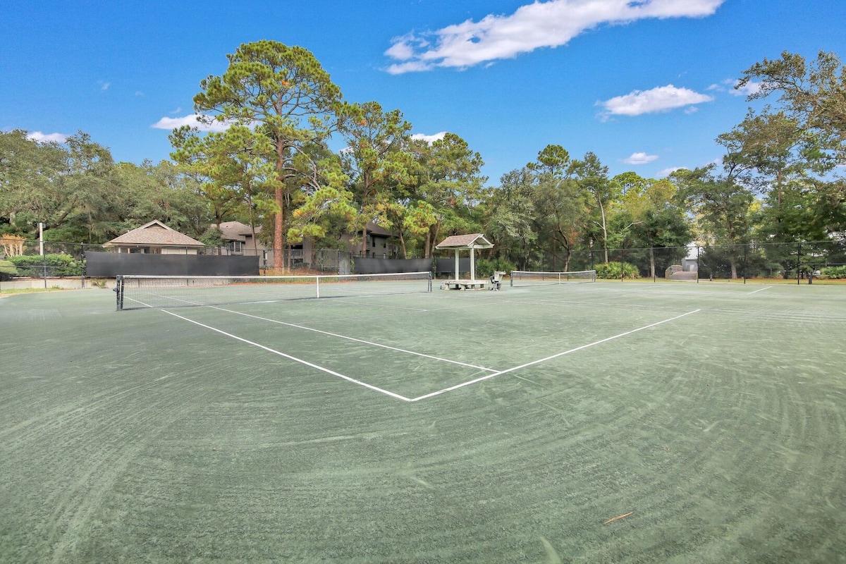 A well-maintained tennis court is set against a backdrop of tall trees and a clear blue sky. The court features marked lines and a net at its center, with a shaded viewing area visible to the side, providing a space for spectators.