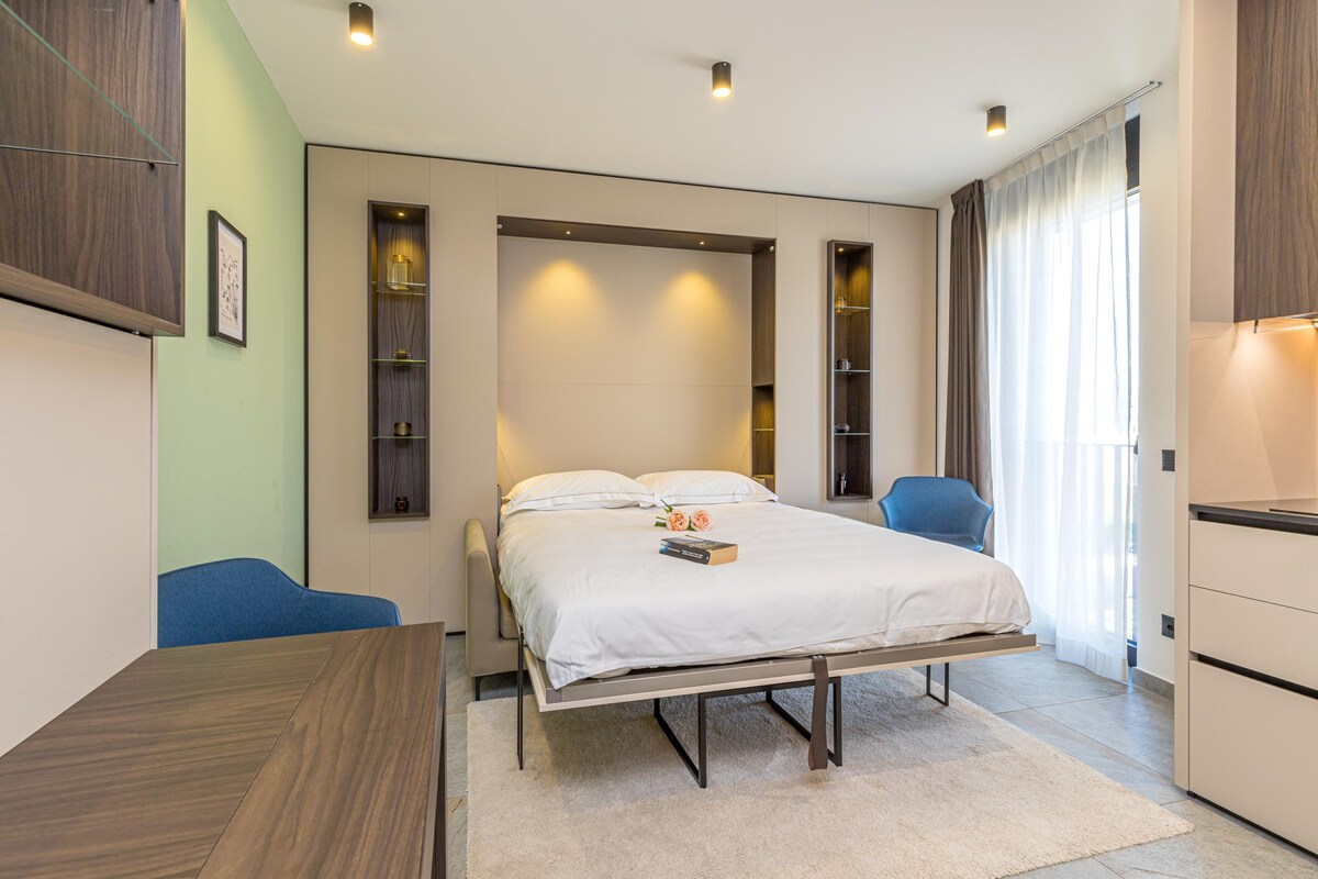A modern studio apartment features a wall bed with a light wood finish, flanked by built-in shelves displaying decorative items. Two contemporary blue chairs are positioned against a wall, with soft lighting enhancing the ambiance. A large window allows natural light to fill the space.