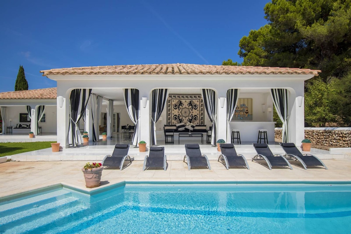 An outdoor terrace is shown, featuring a refreshing pool surrounded by sun loungers. The villa’s elegant façade is highlighted by large open spaces framed with black-and-white striped drapes. Potted plants add a touch of greenery to the sunlit patio area.