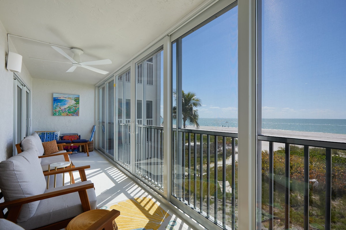 A screened lanai provides a panoramic view of the beach and Gulf waters. Two wooden chairs with cushions are positioned for relaxation, while a vibrant wall hanging adds a touch of color. Sunlight filters through the screens, creating a serene atmosphere.