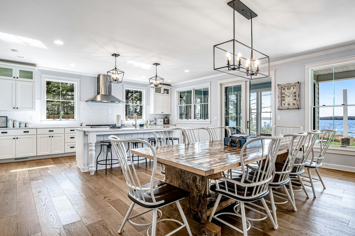 The kitchen and dining area features an open layout with modern white cabinetry and a large farmhouse table that seats up to ten. Pendant lights hang above the table, and large windows offer views of the lake. A spacious kitchen island with bar seating is also visible.