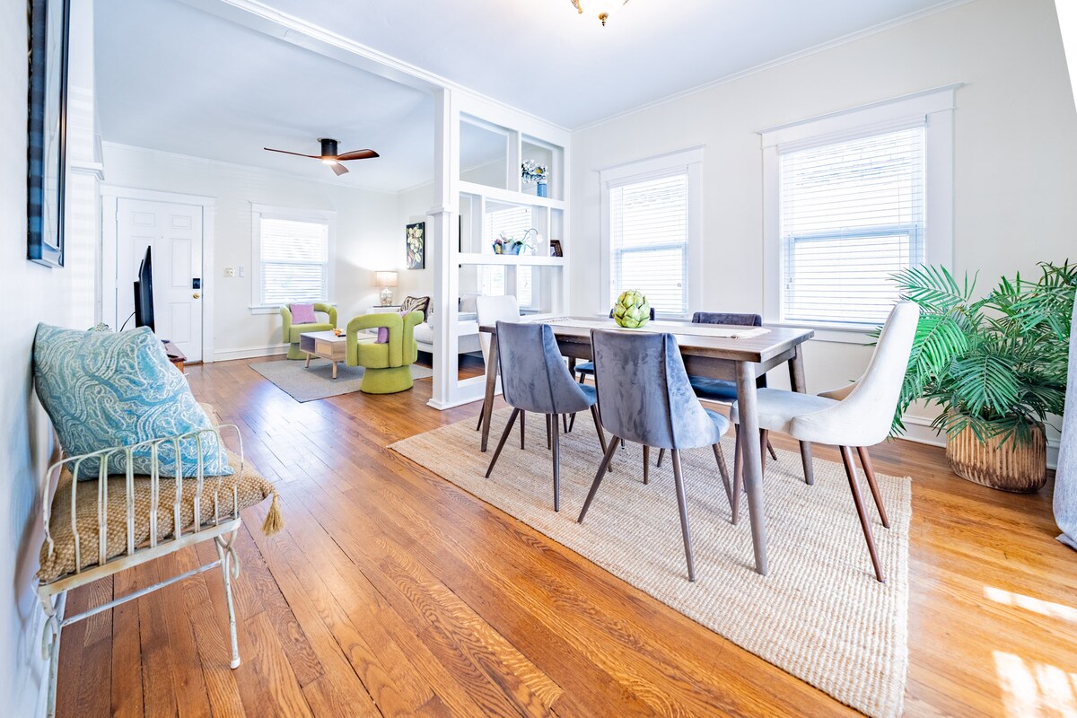 A bright dining area showcases a wooden table surrounded by six upholstered chairs. Natural light fills the space through large windows, highlighting the warm wooden floor. A cozy seating nook is seen in the corner, with a decorative cushion resting on a slender metal bench.