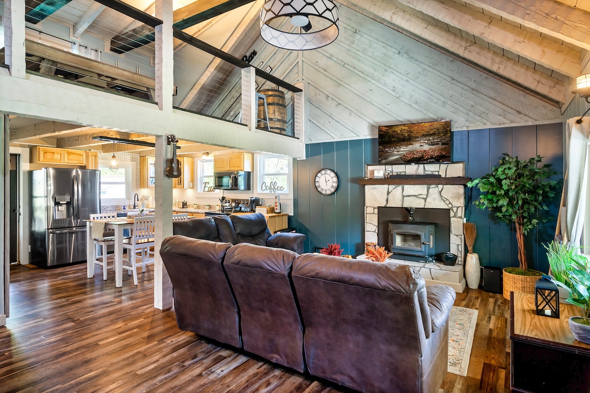 A spacious living area features a comfortable seating arrangement with dark sofas, complemented by a wooden coffee table. The room is enhanced by natural light from large windows, showcasing a charming fireplace and a dining area visible in the background.