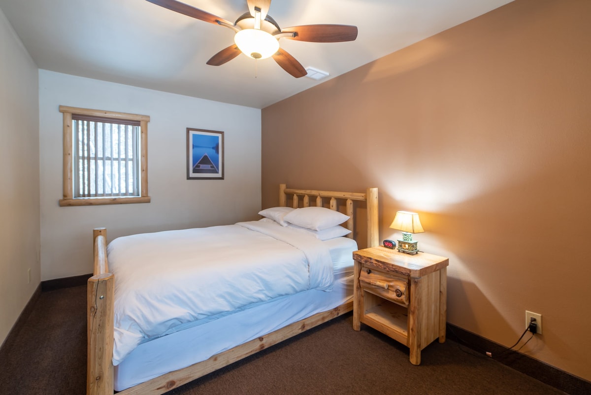 A warm bedroom features a wooden bed with fresh bed linens. A bedside table holds a lamp, providing soft illumination. A window with wooden blinds allows natural light to enter, and a framed artwork adds visual interest to the wall.