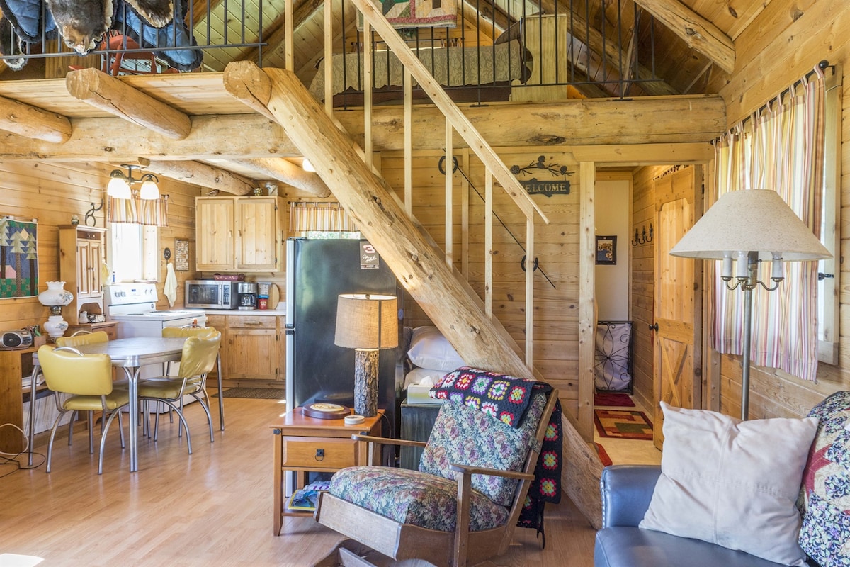 The interior of the cabin features a warm wooden aesthetic, highlighted by log beams and natural light. A staircase leads to the loft bedroom, while a cozy seating area and dining space are arranged thoughtfully. Various amenities, including a refrigerator, are visible within the fully equipped kitchen.
